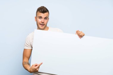 Izole mavi arka plan üzerinde genç yakışıklı sarışın adam eklemek için boş bir beyaz pankart tutarak bir kavram