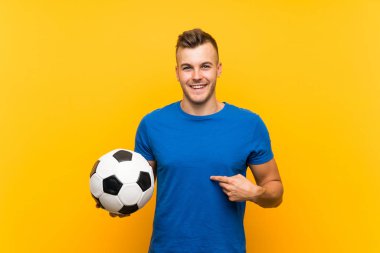 Genç, sarışın bir adam futbol topunu izole edilmiş sarı arka planda tutuyor ve yüzünde sürpriz bir ifade var.