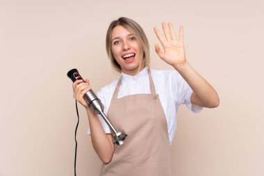 Genç sarışın kadın el blenderi kullanıyor. Mutlu bir ifadeyle selamlıyor.