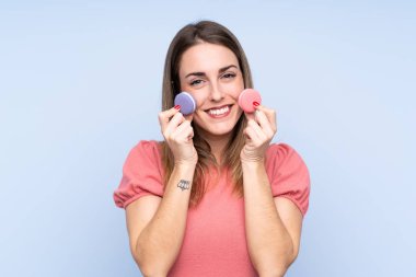 Soyutlanmış mavi arka planda genç sarışın kadın renkli Fransız kurabiyeleri tutuyor ve gülümsüyor.