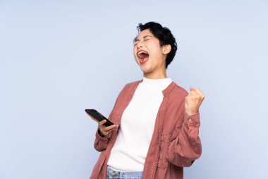 İzole edilmiş mavi arka plan üzerinde genç Asyalı kız ve telefon zafer pozisyonunda.