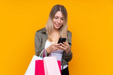 Sarışın, izole edilmiş, sarı arka planı olan genç bir kadın alışveriş torbaları tutuyor ve bir arkadaşına cep telefonuyla bir mesaj yazıyor.