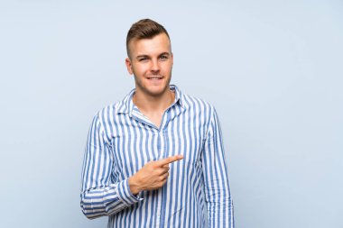 Soyutlanmış mavi arka planı olan yakışıklı sarışın genç bir adam bir ürün sunmak için kenarı işaret ediyor.