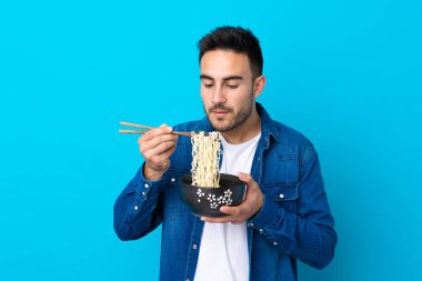 Soyutlanmış mavi arka plan üzerinde genç yakışıklı bir adam elinde çubuklu bir kase erişte tutuyor ve sıcak oldukları için üflüyor.