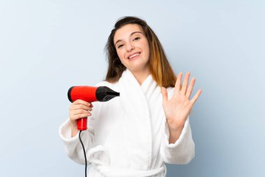 Bornozlu genç bir kadın saç kurutma makinesiyle selamlıyor mutlu bir ifadeyle selamlıyor.