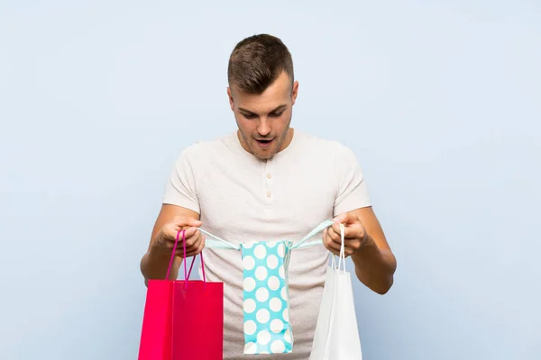 ショッピングバッグの多くを保持する孤立した青い背景の上に若いハンサムなブロンドの男 — ストック写真