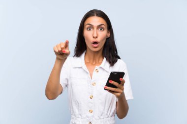İzole edilmiş mavi duvarın üzerinde cep telefonu olan genç bir kadın şaşırmış ve ön tarafı işaret ediyor.