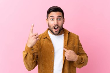 Sakallı genç yakışıklı bir adam pembe arka planda kadife ceket giyiyor ve yüzünde sürpriz bir ifade var.