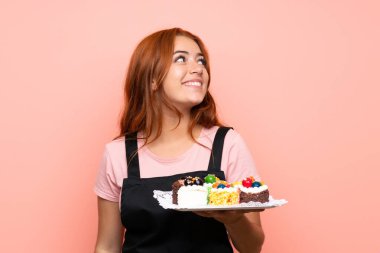 Genç kızıl saçlı kız, elinde bir sürü farklı mini kek tutarak pembe arka plana bakıyor ve gülümsüyor.