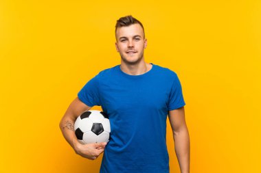 Sarı arka planda futbol topu tutan yakışıklı sarışın genç adam.