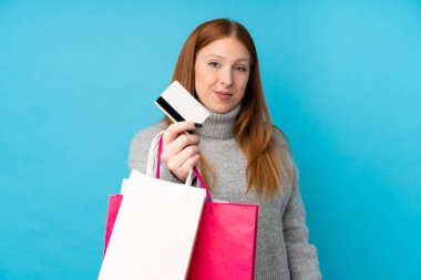 İzole edilmiş mavi arka planı olan genç kızıl saçlı kadın elinde alışveriş çantaları ve bir kredi kartı tutuyor.
