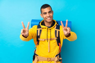 İzole edilmiş mavi arka planda büyük bir sırt çantası olan genç dağcı gülümsüyor ve zafer işareti gösteriyor.