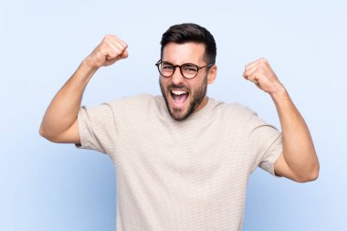 Sakallı yakışıklı genç adam mavi arka planda zaferi kutluyor.