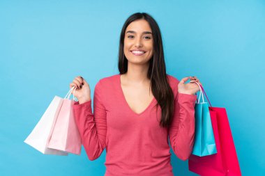 Genç esmer kadın izole edilmiş mavi arka planda alışveriş torbaları tutuyor ve gülümsüyor.