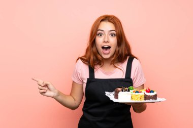Genç kızıl saçlı kız, izole edilmiş pembe arka planda bir sürü farklı mini kek tutuyor. Şaşırmış ve işaret ediyor.