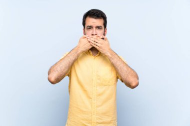 İzole edilmiş mavi arka planda sakallı genç bir adam elleriyle ağzını kapatıyor.