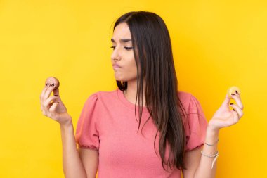 Genç esmer kadın izole edilmiş sarı arka planda renkli Fransız kurabiyeleri tutuyor ve mutsuz.