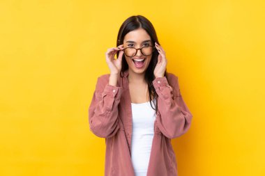 Genç esmer kadın, izole edilmiş sarı arka planda, gözlüklü ve şaşırmış.