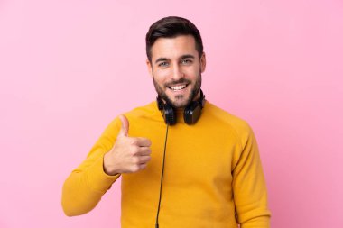 Kulaklıkları olan genç yakışıklı bir adam pembe arka planda. Başparmakları havada. Çünkü iyi bir şey oldu.