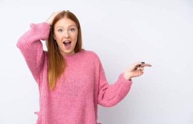 İzole edilmiş beyaz arka planda pembe kazaklı genç, kızıl saçlı kadın şaşırmış ve parmağını kenarı işaret ediyor.