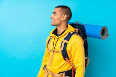 İzole edilmiş mavi arka plan üzerinde büyük bir sırt çantası olan genç dağcı yan tarafa bakıyor.