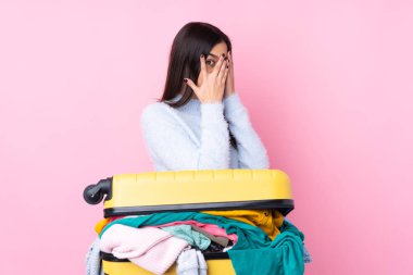 Bir bavul dolusu elbisesi olan gezgin kadın izole edilmiş pembe arka planda gözleri kapatıyor ve parmaklarından bakıyor.