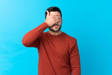 Sakallı, genç, yakışıklı, soyutlanmış, mavi arka planda, gözleri elle kaplanmış.