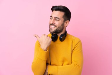 İzole edilmiş pembe arka planda kulaklıkları olan genç yakışıklı bir adam bir ürün sunmak için kenarı işaret ediyor.