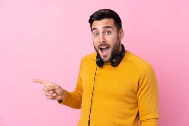 Soyutlanmış pembe arka planda kulaklıkları olan genç ve yakışıklı bir adam şaşırmış ve parmağıyla kenarı işaret ediyor.