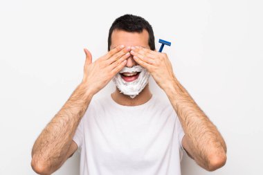 Sakalını kesen bir adam izole edilmiş beyaz arka planda gözlerini elleriyle kapatıyor.