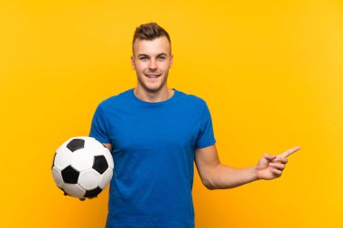 Sarı arka planda futbol topu tutan yakışıklı sarışın genç adam şaşırdı ve işaret etti.
