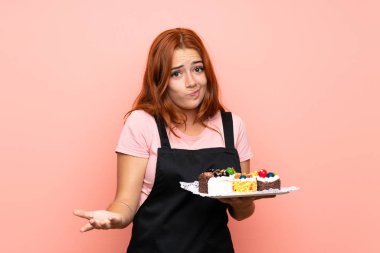 Genç kızıl saçlı kız pembe arka planda bir sürü farklı mini kek tutuyor. Omuzlarını kaldırırken şüphe uyandırıyor.
