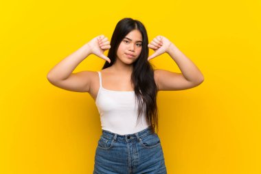 Genç Asyalı kız izole edilmiş sarı arka plan başparmağını aşağı gösteriyor.