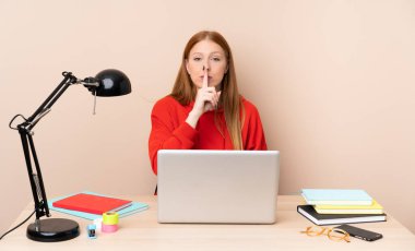 İşyerinde dizüstü bilgisayarı olan genç bir bayan sessiz bir jest gösteriyor. Parmağını ağzına sokuyor.