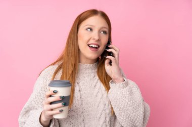 Genç kızıl saçlı, soyutlanmış pembe arka planda, elinde kahve ve cep telefonu olan bir kadın.