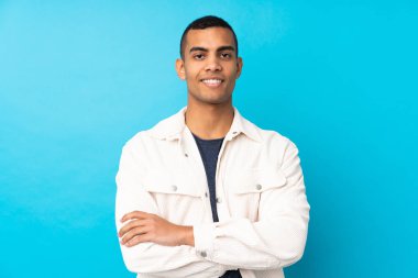 Genç Afro-Amerikalı adam mavi arka planda kollarını ön cephede çapraz tutuyor.