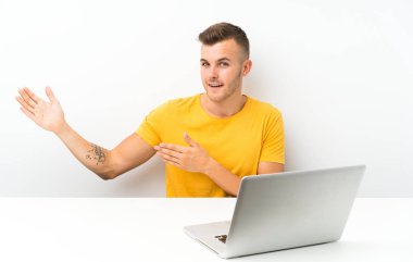 Masada dizüstü bilgisayarı olan genç sarışın bir adam davet edildiği için ellerini yana uzatıyor.