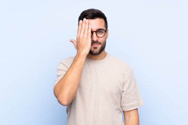 Sakallı, genç ve yakışıklı bir adam. Tek başına mavi arka planda. Gözünü kapatıyor.