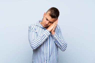 Genç, sarışın, soyutlanmış, mavi arka planda bir adam uyku jesti yapıyor.