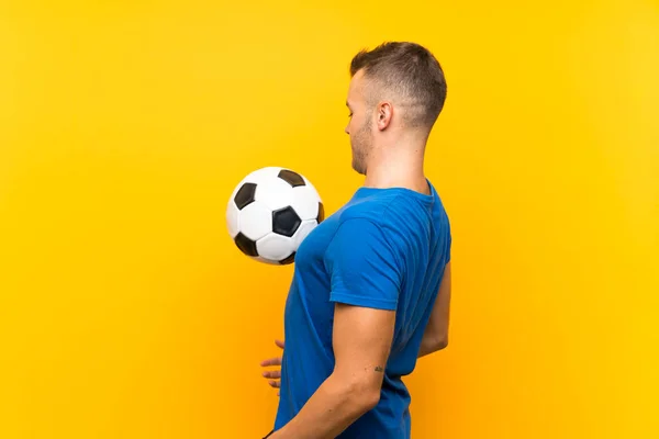 Jovem homem loiro bonito segurando uma bola de futebol sobre fundo amarelo isolado — Fotografia de Stock