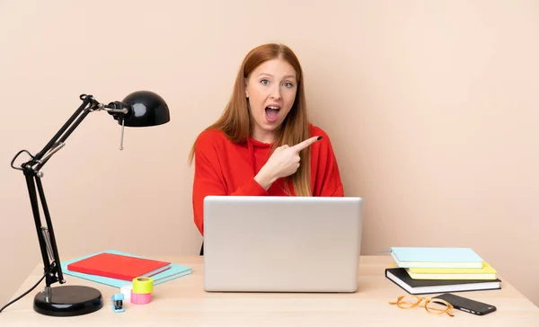 Young student woman in a workplace with a laptop surprised and pointing side