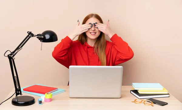 Young student woman in a workplace with a laptop covering eyes by hands