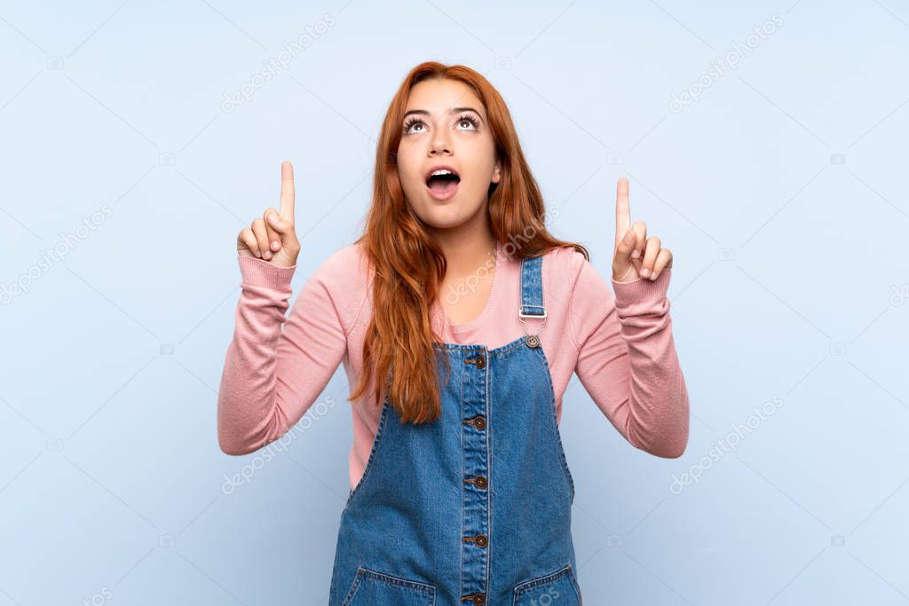 Teenager redhead girl with overalls over isolated blue background pointing with the index finger a great idea