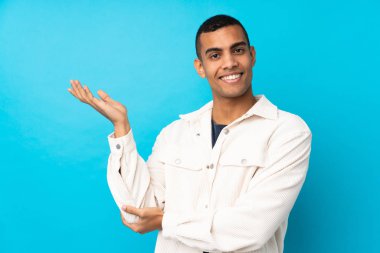 İzole edilmiş mavi arka planda yaşayan genç bir Afro-Amerikan erkeği davet etmek için ellerini yana uzatıyor.
