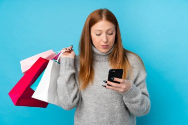 Genç kızıl saçlı kadın, mavi arka planda, elinde alışveriş çantaları ve cep telefonuyla bir arkadaşına mesaj yazıyor.