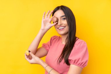 Genç esmer kadın izole edilmiş sarı arka planda elinde renkli Fransız kurabiyeleri tutuyor.