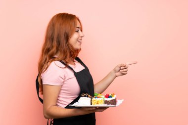 İzole edilmiş pembe arka planda bir sürü farklı mini kek tutan genç kızıl kız bir ürünü sunmak için kenarı işaret ediyor.