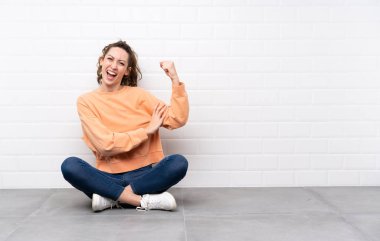 Yerde oturan kıvırcık saçlı genç kadın güçlü bir jest yapıyor.