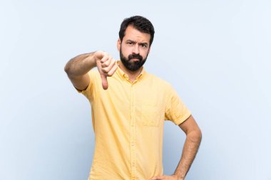 İzole edilmiş mavi arka planda sakallı genç bir adam olumsuz bir ifadeyle baş parmağını gösteriyor.