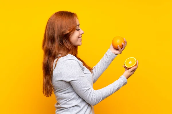 Teenager redhead girl holding an orange over isolated yellow background — Stock Photo, Image